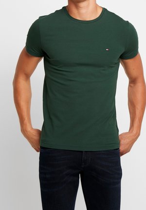 Man wearing dark green fitted short-sleeve T-shirt with small logo on chest and dark blue jeans, hands positioned near pockets.