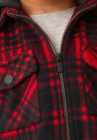 Röd och svart rutig fleecjacka med dragkedja fram, två bröstfickor och grå huva. Strukturerad stof med en mysig utseende.