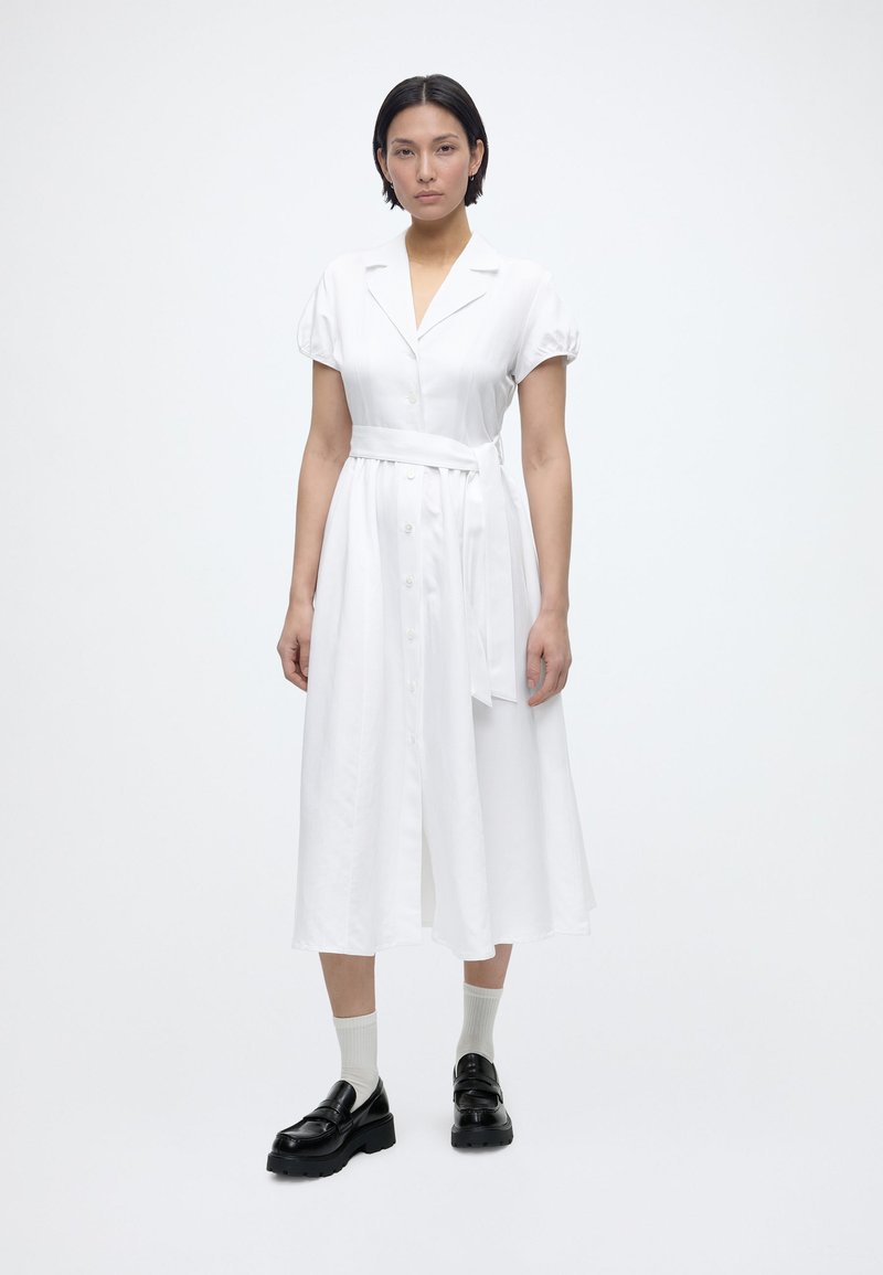 Γυναίκα που φοράει λευκό midi φόρεμα με κουμπιά, κοντά μανίκια και ζώνη, σετ με λευκές κάλτσες και μαύρα χοντρά μοκασίνια.