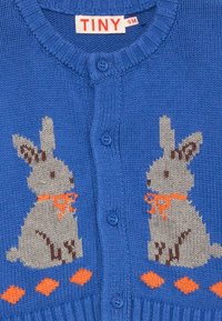Cardigã azul de malha com dois coelhos bege com pormenores em laranja. Terminado com botões azuis e uma bainha canelada. Etiqueta de tamanho: PEQUENO, 9M.