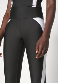 Leggings atléticos negros con paneles laterales blancos, tejido elástico, cinturilla alta y costuras planas, que ofrecen una silueta ajustada.