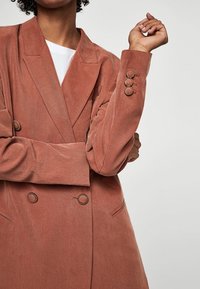 Blazer doppiopetto in terracotta con tessuto texture, rever a rovescio, dettaglio con tre bottoni sui polsini e tasche frontali, indossato sopra una maglietta bianca.