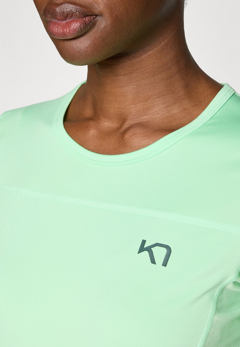 Jasnozielona koszulka sportowa z okrągłym dekoltem, z ciemnym logo po lewej stronie. Gładka tekstura i minimalne szczegóły szycia.