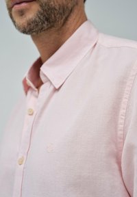Camisa de botones de color rosa claro con cuello, que presenta una tela texturizada y un sutil logotipo bordado en el pecho.