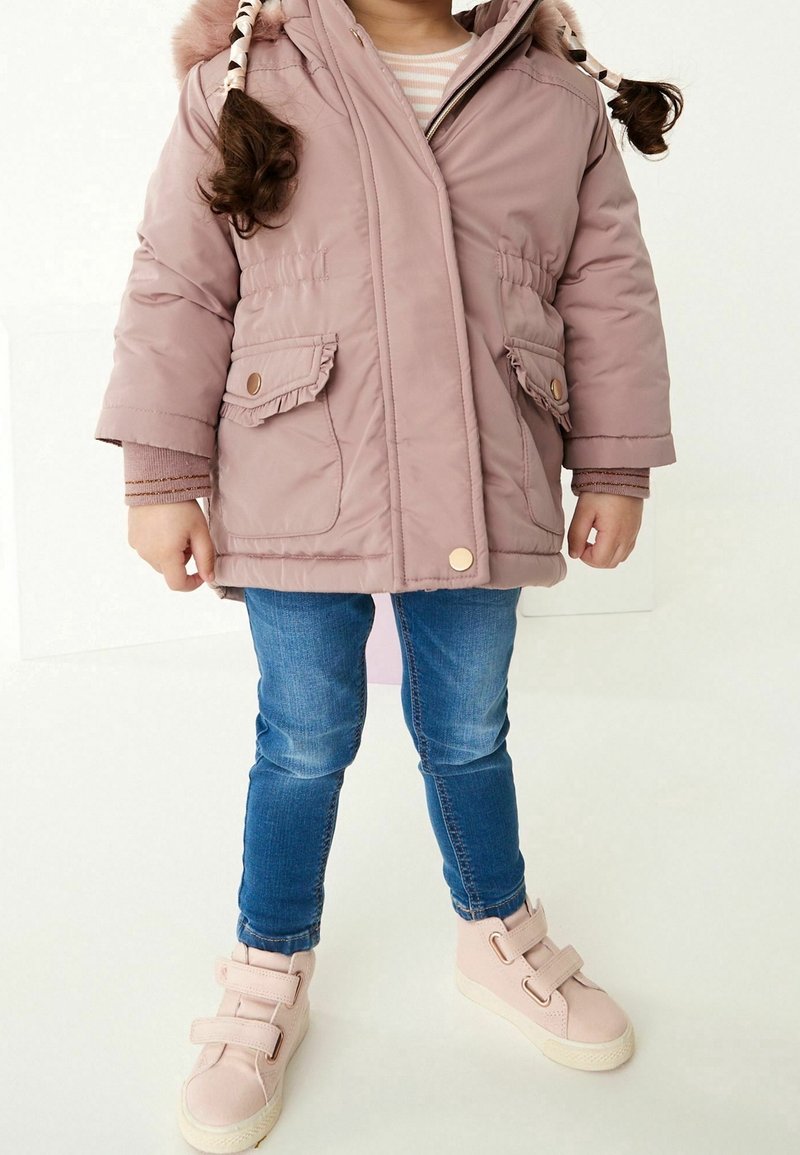 Enfant aux cheveux tressés portant une veste d'hiver rose, un jean bleu et des baskets roses à scratch, debout sur un sol blanc devant un fond blanc.
