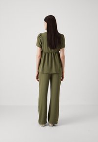 Olivno zelena majica s kratkimi rokavi in peplum pasom, kombinirana z širokimi hlačami. Mehka textura; minimalističen dizajn. Stoječa figura, pogled od zadaj.