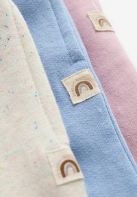 Tre tygprover i beige med färgglada prickar, ljusblå och rosa. Varje har en liten vävd etikett med en brun regnbågsdesign.