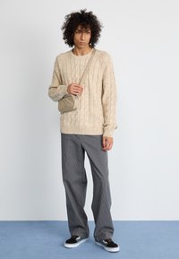 Bež pulover z pletenim vzorcem, sivi hlače s črtami, običajni športni čevlji. Model nosi majhno bež torbico čez ramo in stoji na modri površini.