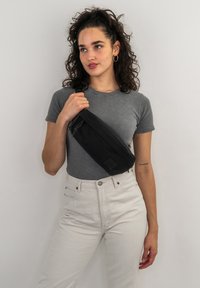 Borsa a spalla nera in tela con un design elegante, hardware discreto e un pannello con il logo, indossata da una persona con una t-shirt grigia e pantaloni chiari.