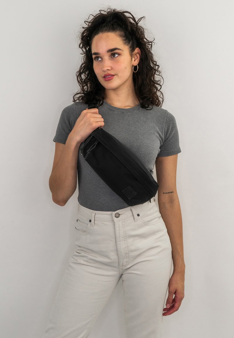 Borsa a spalla nera in tela con un design elegante, hardware discreto e un pannello con il logo, indossata da una persona con una t-shirt grigia e pantaloni chiari.