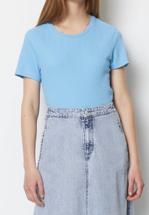 Vrouw draagt een lichtblauw geribd T-shirt met korte mouwen, ingestopt in een lichtblauwe spijkerrok met hoge taille, voorzien van knoop- en ritssluiting vooraan.