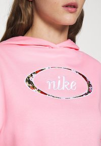 Rosa hoodie med en mjuk textur, med en cirkulär logotyp med blommotiv och texten "nike" i vitt, samt en justerbar huva.