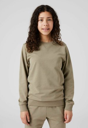 Ung person med krøllet mørkt hår iført en løs, olivengrøn sweatshirt og matchende bukser, stående mod en ensfarvet lys baggrund.