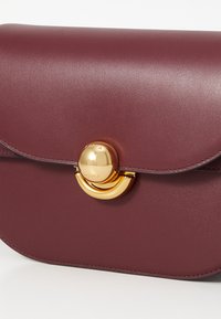 Borsa in pelle bordeaux con finitura liscia, patta arrotondata e dettaglio della chiusura circolare in tonalità oro. Forma compatta e strutturata.