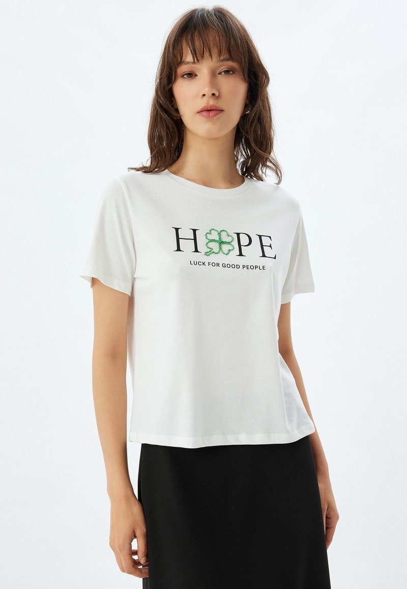 Hvid bomulds T-shirt med korte ærmer. Har "HOPE" i sort tekst og en grøn firkløver, med "LUCK FOR GOOD PEOPLE" nedenunder.