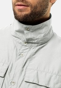 Camicia grigia chiara con colletto alto, dotata di chiusura frontale a bottoni e due tasche sul petto. Il tessuto appare liscio con una leggera lucentezza.