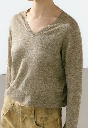 Personne portant un pull beige à col en V et un pantalon en daim marron clair, avec un fond clair uni.