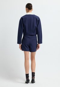 Granatowy dwuczęściowy strój z krótką kurtką z białym kołnierzem i dopasowanymi szortami, połączony z czarnymi szpilkami i czarnymi skarpetkami.