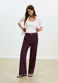 Pantaloni larghi di colore bordeaux realizzati in tessuto testurizzato, dotati di una cintura nera con fibbia argentata. Indossati con una camicia bianca e una canotta.