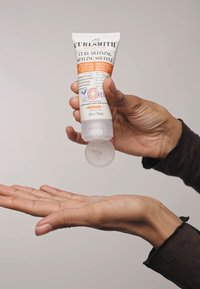 Curlsmith Curl Defining Styling Soufflé, witte tube met oranje accenten, 2 fl oz, gel uitknijpend op een handpalm; gladde textuur, licht reflectie.