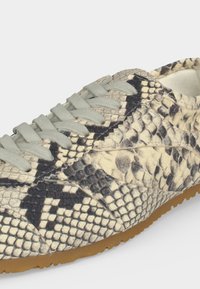 Baskets basses avec motif peau de serpent beige et noir, lacets gris clair et semelle en caoutchouc marron clair sur fond blanc.