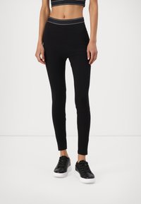 Leggings noirs avec une taille haute arborant une bande logo subtile. Associés à des baskets noires et un crop top assorti. Texture lisse.