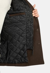 Giacca esterna marrone con interno trapuntato nero. Presenta una fodera grigia testurizzata, chiusura con cerniera e una piccola tasca interna.