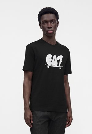 Νέος άνδρας που φοράει μαύρο t-shirt με λευκό λογότυπο "EA7" και γραφικό σκέιτμπορντ, στέκεται μπροστά σε απλό φωτεινό φόντο.