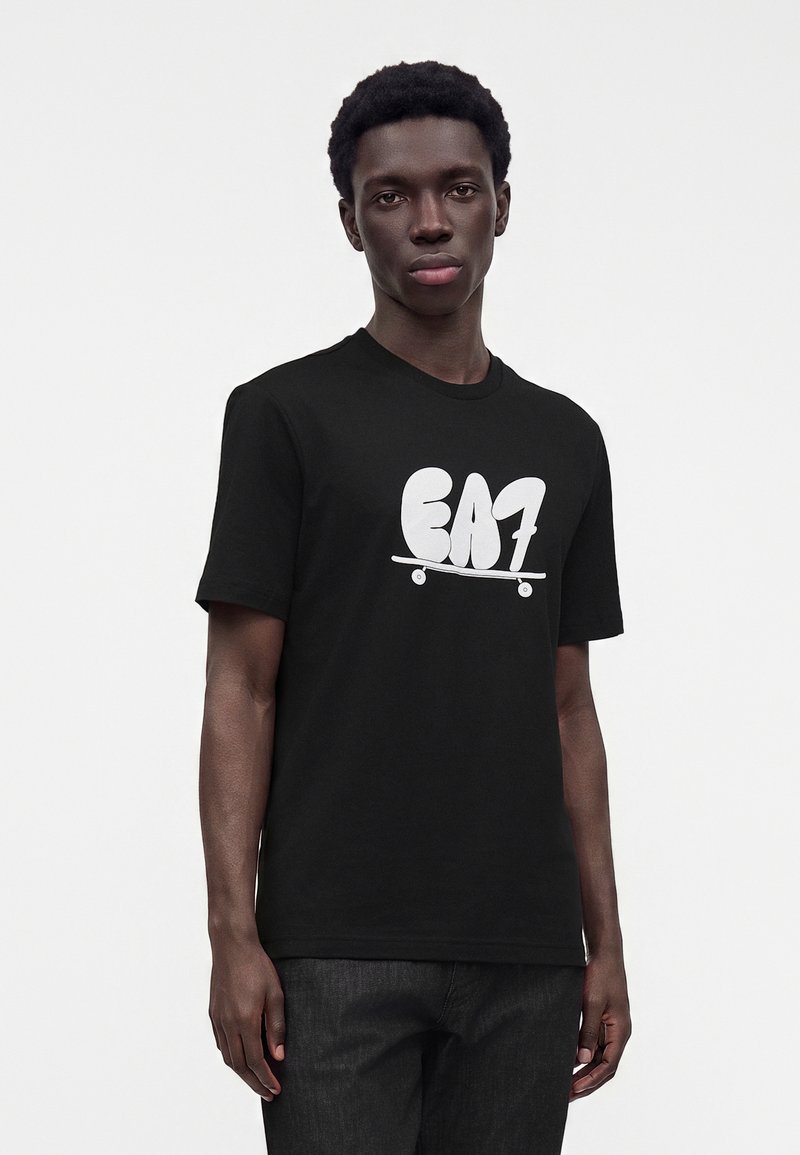 Νέος άνδρας που φοράει μαύρο t-shirt με λευκό λογότυπο "EA7" και γραφικό σκέιτμπορντ, στέκεται μπροστά σε απλό φωτεινό φόντο.