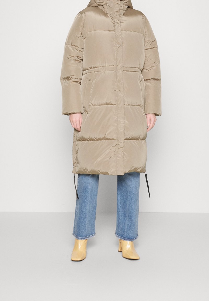 Manteau long matelassé beige avec capuche, poches avant, porté sur un jean bleu et des bottines jaunes à talons blocs.