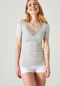 Top gris de manga corta con un escote en V adornado con encaje y un patrón texturizado. La modelo lo combina con pantalones cortos blancos, destacando el ajuste de la camisa.