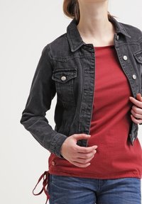 Svart denimjacka med två bröstfickor, silverdetaljer och en åtsittande design, lager-på-lager över en röd strukturerad topp och tillsammans med blå jeans.