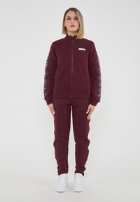 Burgundowa bluza z zamkiem i wysokim kołnierzem, z logo na piersi i graficznymi akcentami na rękawach, w połączeniu z pasującymi spodniami dresowymi i białymi butami sportowymi.