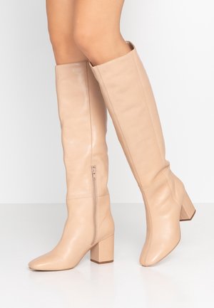 Beiget nahkaiset polven korkeudelle yltävät saappaat, joissa on paksut korot ja sivuvetoketjut, jalassa valkoista taustaa vasten.