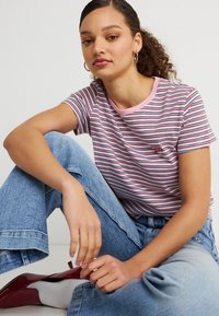 Νεαρή γυναίκα με σγουρά μαλλιά που φοράει κρίκους στα αυτιά, ριγωτό μπλουζάκι, ανοιχτόχρωμο τζιν, λευκές κάλτσες και κόκκινα παπούτσια καθισμένη με σταυρωμένα πόδια μπροστά σε λευκό φόντο.