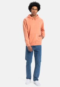 Sudadera color durazno con cordón, bolsillo tipo canguro y puños acanalados. Combinada con jeans azules y zapatillas blancas sin cordones.