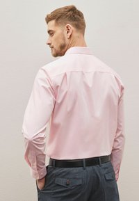 Helles rosa Langarmhemd mit spitzem Kragen, geknöpften Manschetten und Rückennahtfalten; kombiniert mit dunklen strukturierten Hosen und einem schwarzen Gürtel.