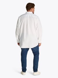 Camisa blanca de manga larga con un ajuste relajado, dobladillo curvado y cuello abotonado, combinada con unos vaqueros de mezclilla azul oscuro y zapatillas blancas.