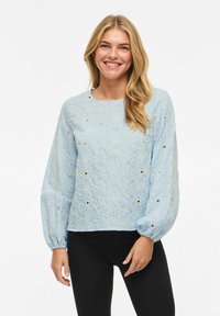 Femme souriante aux longs cheveux blonds portant un chemisier bleu clair à motifs floraux ajourés avec des manches bouffantes et un pantalon noir, debout devant un fond blanc.