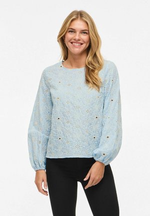 Femme souriante aux longs cheveux blonds portant un chemisier bleu clair à motifs floraux ajourés avec des manches bouffantes et un pantalon noir, debout devant un fond blanc.