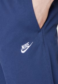 Pantalons de survêtement navy en tissu doux, dotés d'une poche incurvée et d'un logo Nike blanc brodé sur le côté. Texture lisse avec un design simple.