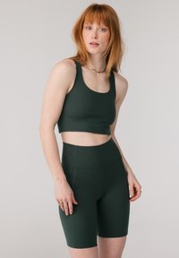 Kvinne med rødt hår iført mørkegrønn sport-BH og matchende høytlivs biker-shorts mot en ensfarget lys bakgrunn.