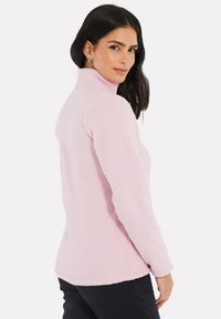Veste en polaire rose clair avec col haut, manches longues et surface texturée. Présente un ourlet légèrement arrondi à l'arrière.