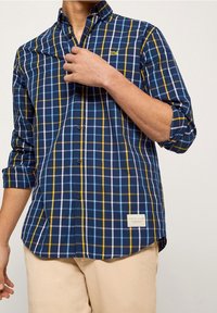 Modrá kostkovaná košile se žlutými a bílými akcenty, dlouhé rukávy s ohrnutím k loktům, knoflíková léga a štítek na dolním pravém rohu.