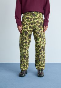 Camo joggings in groen, bruin en zwart, met een hoge taille, losse pasvorm en grote zakken, gecombineerd met zwarte schoenen met groene accenten.