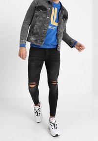 Jeans negros ajustados con rodilleras rasgadas, combinados con un sudadera azul con letras amarillas y una chaqueta de denim descolorida. Zapatillas blancas.