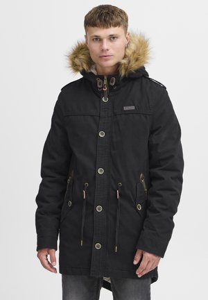 Parka negra con capucha de piel sintética, cremalleras y botones en la parte frontal, cintura con cordón ajustable y bolsillos laterales con detalles en bronce.