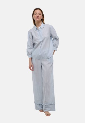 Femme portant un haut de pyjama rayé bleu clair à manches longues avec un col volanté et un pantalon large assorti avec bordure à volants, pieds nus, debout.