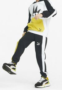 Czarny i biały hoodie z żółtymi akcentami i kreskówkowym wzorem, w połączeniu z czarnymi spodniami dresowymi z bocznymi paskami i logo. Czarne sneakersy z kwiatowym wzorem.