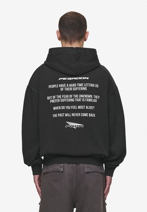 Homme portant un sweat à capuche noir avec un texte blanc au dos : phrases motivantes sur la souffrance, la peur et la vie, en police minimaliste.